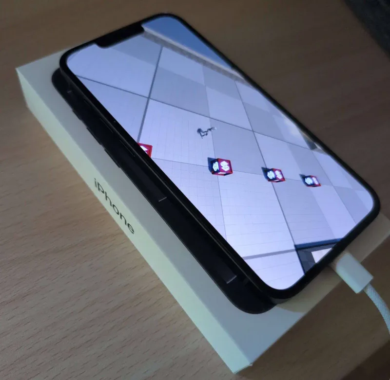 An iPhone lying on its box, plugged in and running a Stride sample scene with a small character and red crates on a grey grid floor.